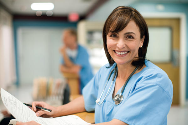 Female doctor wearing blue uniform and a stethoscope around her neck. Holding pen and paper. Smiling.